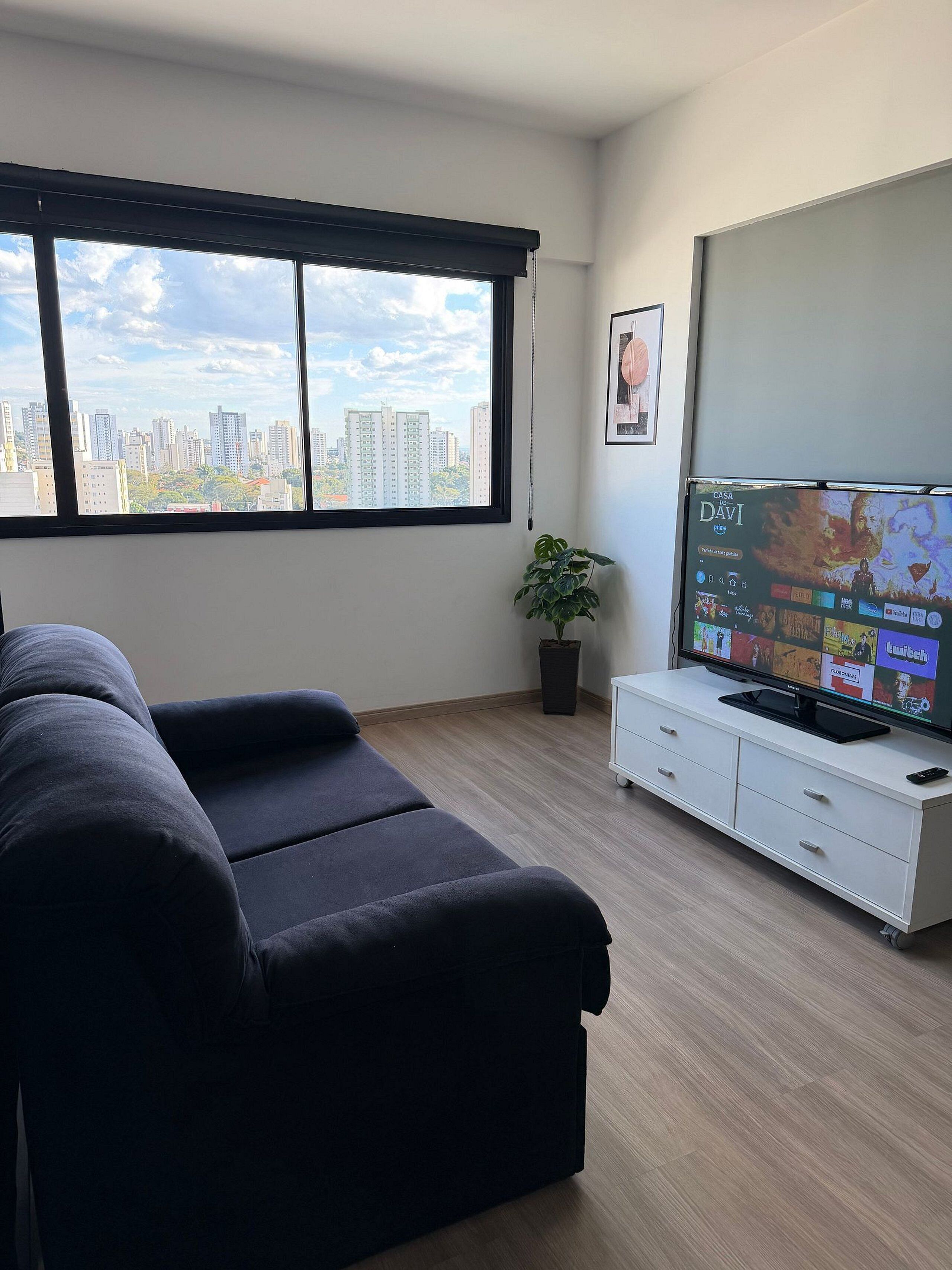 Sala de estar moderna com sofá, televisão exibindo um filme, planta em vaso e vista da cidade através de grandes janelas.