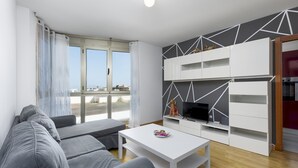 Een moderne woonkamer met een bank, een salontafel en een televisie op een witte kast. Er is een groot raam met gordijnen, en een keuken met witte kasten en een rode apparatuur.