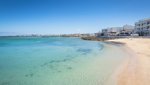Een strand met helder turquoise water, een zandrijk strand en witte gebouwen op de achtergrond.
