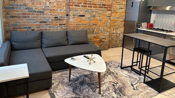 A modern living room with a grey sectional sofa, a white coffee table, and a marble-patterned rug. A kitchen area with a stainless steel refrigerator and a gas stove is visible in the background. - Cozy 1-bedroom apartment in brilliant Cincinnati with AC (Cincinnati)
