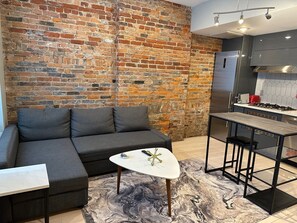 Un salon moderne avec un canapé d’angle gris, une table basse blanche et un tapis à motifs de marbre. On aperçoit à l’arrière-plan un coin cuisine avec un réfrigérateur en acier inoxydable et une cuisinière à gaz.