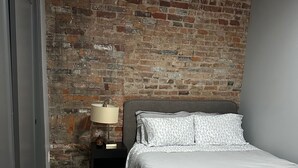 A bedroom with a brick wall, a bed with white bedding, a nightstand with a lamp, and a patterned rug. - Cozy 1-bedroom apartment in brilliant Cincinnati with AC (Cincinnati)