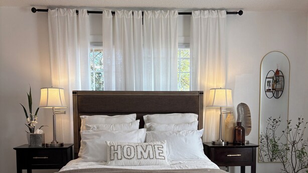 A bedroom with a large bed, two bedside tables with lamps, a window with sheer curtains, and a wall-mounted mirror.