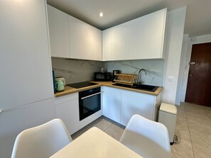 A compact kitchen with white cabinets, a black countertop, a sink, a stove, and a microwave. There are two white chairs and a small trash bin. - Cozy apartment with terrace in Cannes (Cannes)