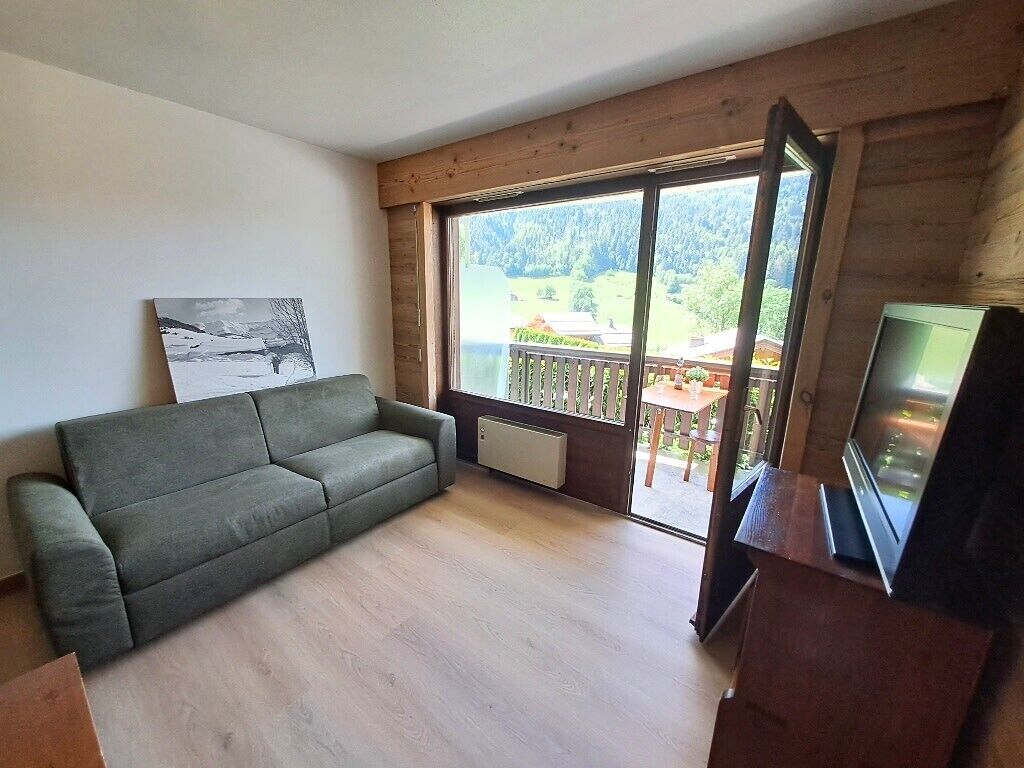 Une pièce comprenant un canapé, un téléviseur, un tableau et une grande fenêtre donnant sur un balcon et de la verdure.