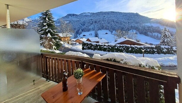 Un balcón con una mesa de madera y un vaso de agua, con vistas a un paisaje nevado con casas y árboles.