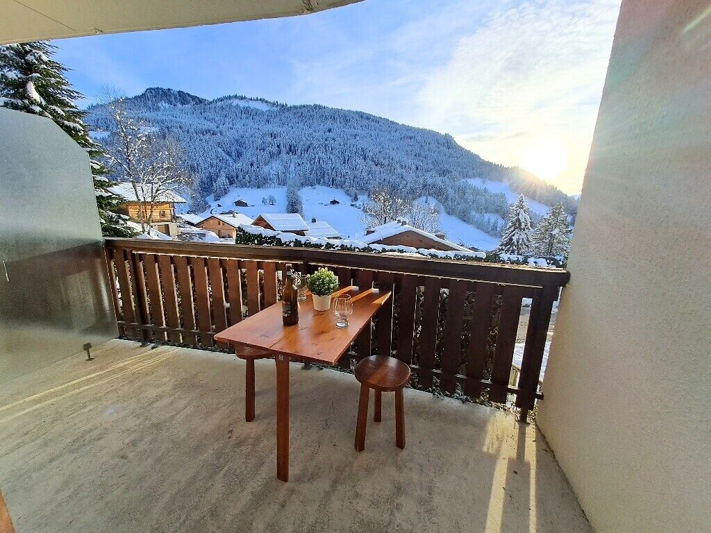 A balcony with a wooden table and chairs, overlooking a snow-covered mountain and houses.