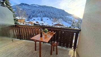Un balcon avec une table et des chaises en bois, donnant sur une montagne enneigée et des maisons.