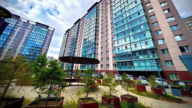A modern high-rise building with multiple floors, large windows, and a parking area with cars and greenery in the foreground.