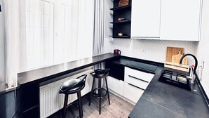 A modern kitchen with black countertops, white cabinets, and a black backsplash. There are two bar stools and a wooden cutting board on the countertop.