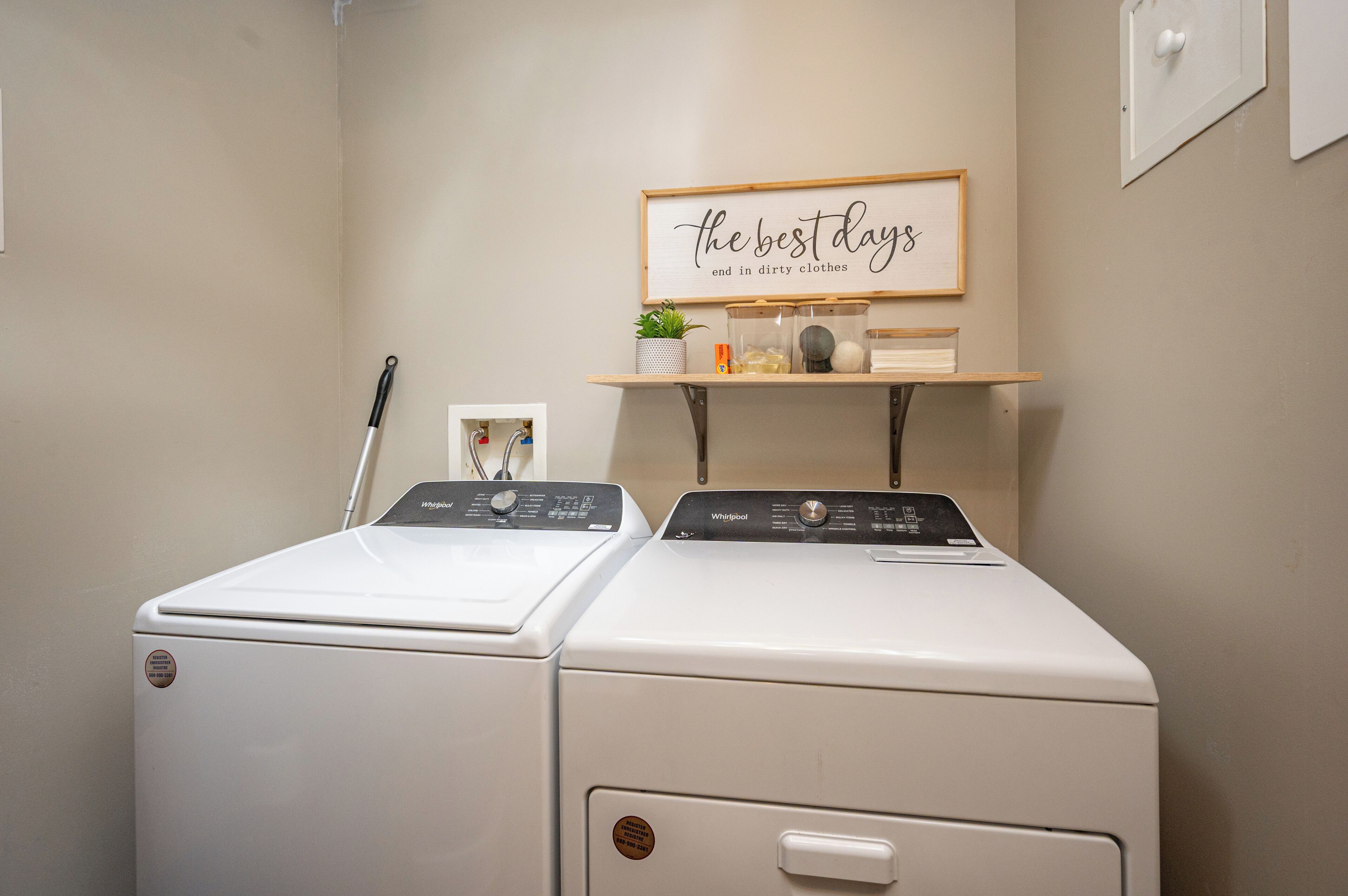 A laundry room with two stacked washers and dryers, a shelf with a potted plant, and a framed sign on the wall.