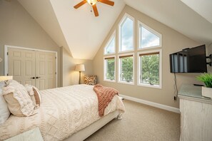 Ein Schlafzimmer mit einem großen Bett, einem Deckenventilator, einem Fernseher und einem Fenster mit Blick auf Bäume.