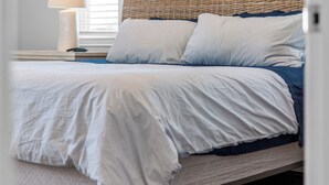 Una cama bien hecha con edredón blanco y ropa de cama azul, cabecero de mimbre y una lámpara de noche.