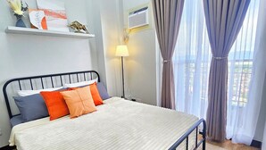 A neatly made bed with a white comforter and multiple pillows, a bedside lamp, a window with curtains, and a small shelf with decorative items. - Condo in Davao City - 2BR Penthouse @ Verdon Parc (Davao City)