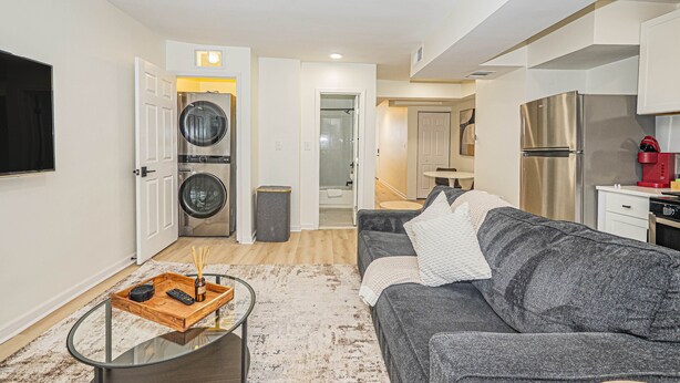 Una sala de estar moderna con un sofá gris, una mesa de centro de vidrio y un televisor. Hay un área de lavandería con lavadora y secadora.