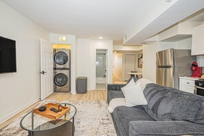Ein modernes Wohnzimmer mit einem grauen Sofa, einem Glastisch und einem Fernseher. Es gibt eine Waschküche mit Waschmaschine und Trockner.