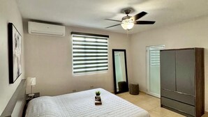 Ein Schlafzimmer mit Bett, Deckenventilator, Klimaanlage, Fenster mit Jalousien, Spiegel, Kleiderschrank und einer kleinen Pflanze auf dem Bett.