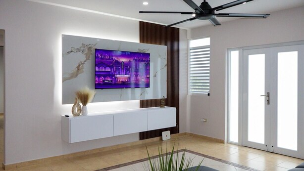 Ein modernes Wohnzimmer mit Wand-Fernseher, Deckenventilator und einem großen Fenster mit Jalousien.