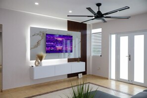 Een moderne woonkamer met een tv aan de muur, een plafondventilator en een groot raam met zonwering.