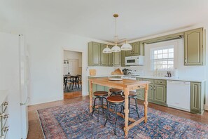 Une cuisine avec des meubles de rangement verts, un îlot central en bois avec des tabourets, un luminaire suspendu et un tapis à motifs.