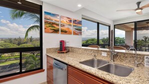 A kitchen with granite countertops, stainless steel sink, and a view of a lush landscape through large windows. - Perfect for a couples getaway!  311 Shores at Waikoloa (Waikoloa Beach Resort)