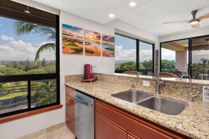 A kitchen with granite countertops, stainless steel sink, and a view of a lush landscape through large windows. - Perfect for a couples getaway!  311 Shores at Waikoloa (Waikoloa Beach Resort)