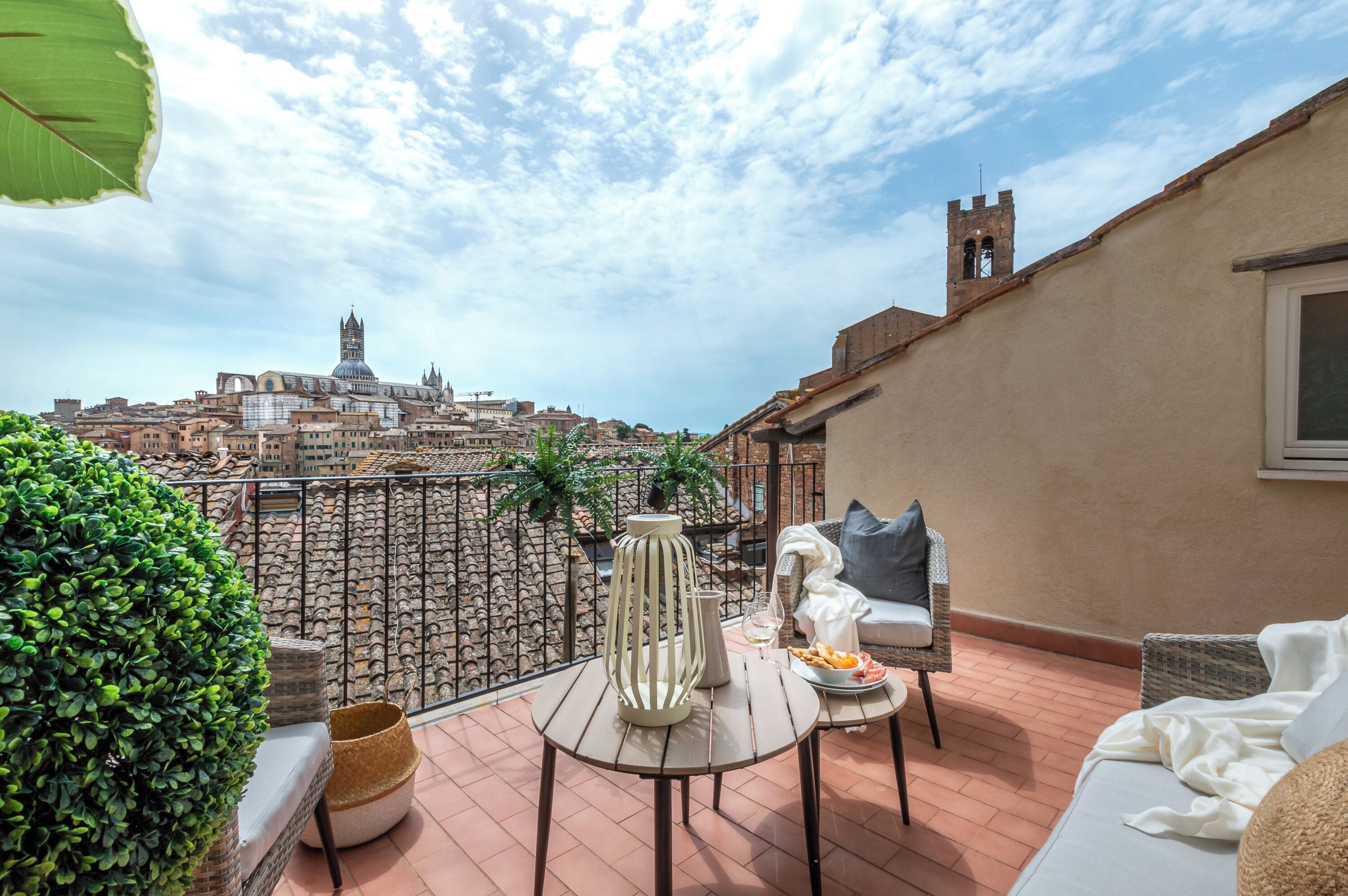 A rooftop terrace with a view of historic buildings, a table with a vase and a plate of food, and a comfortable seating area.