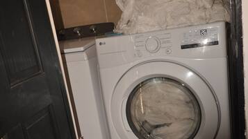 A compact, white washing machine in a small laundry room with a tiled floor and a dark door.