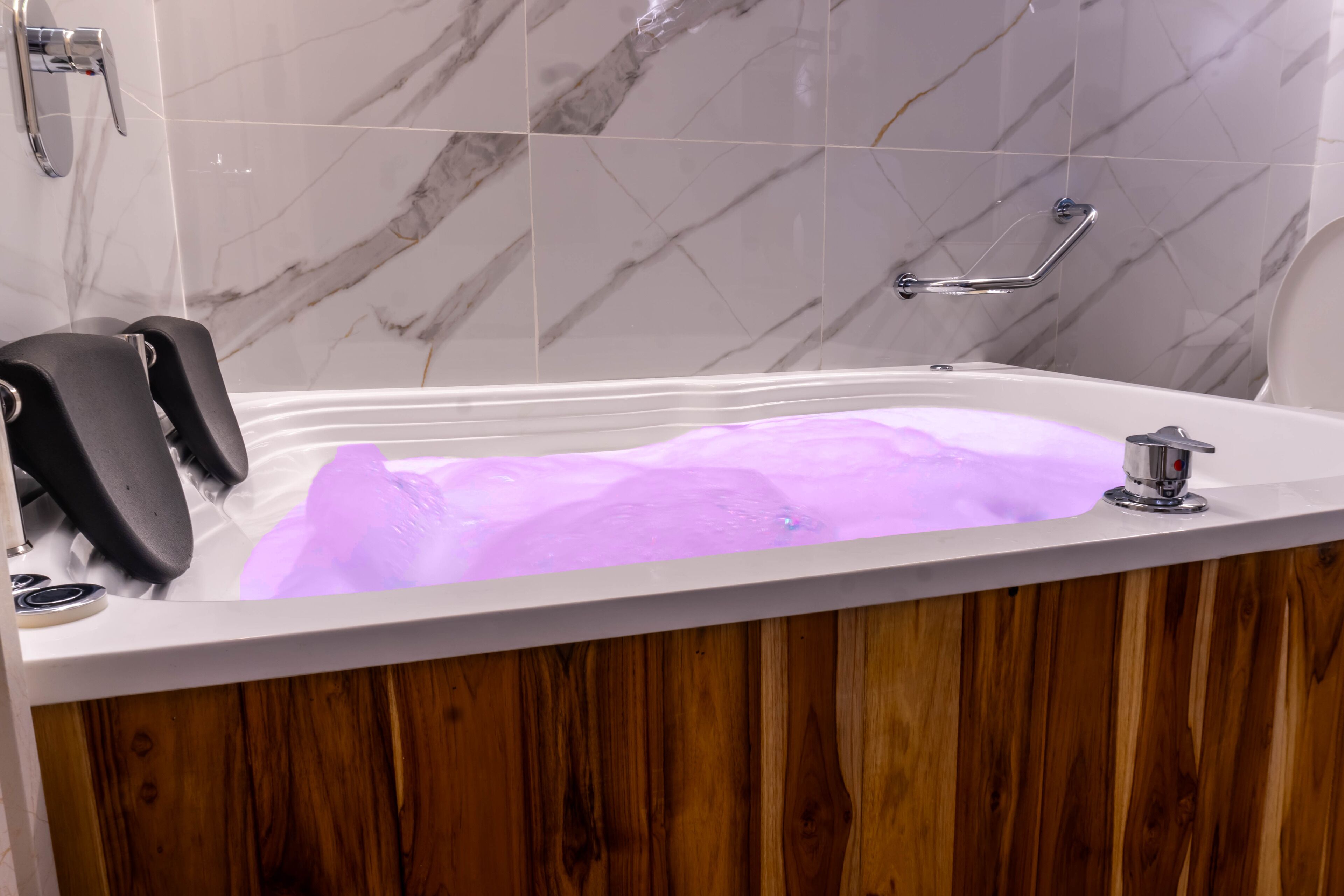 A bathtub with a wooden panel, a handrail, and a marble-tiled wall.