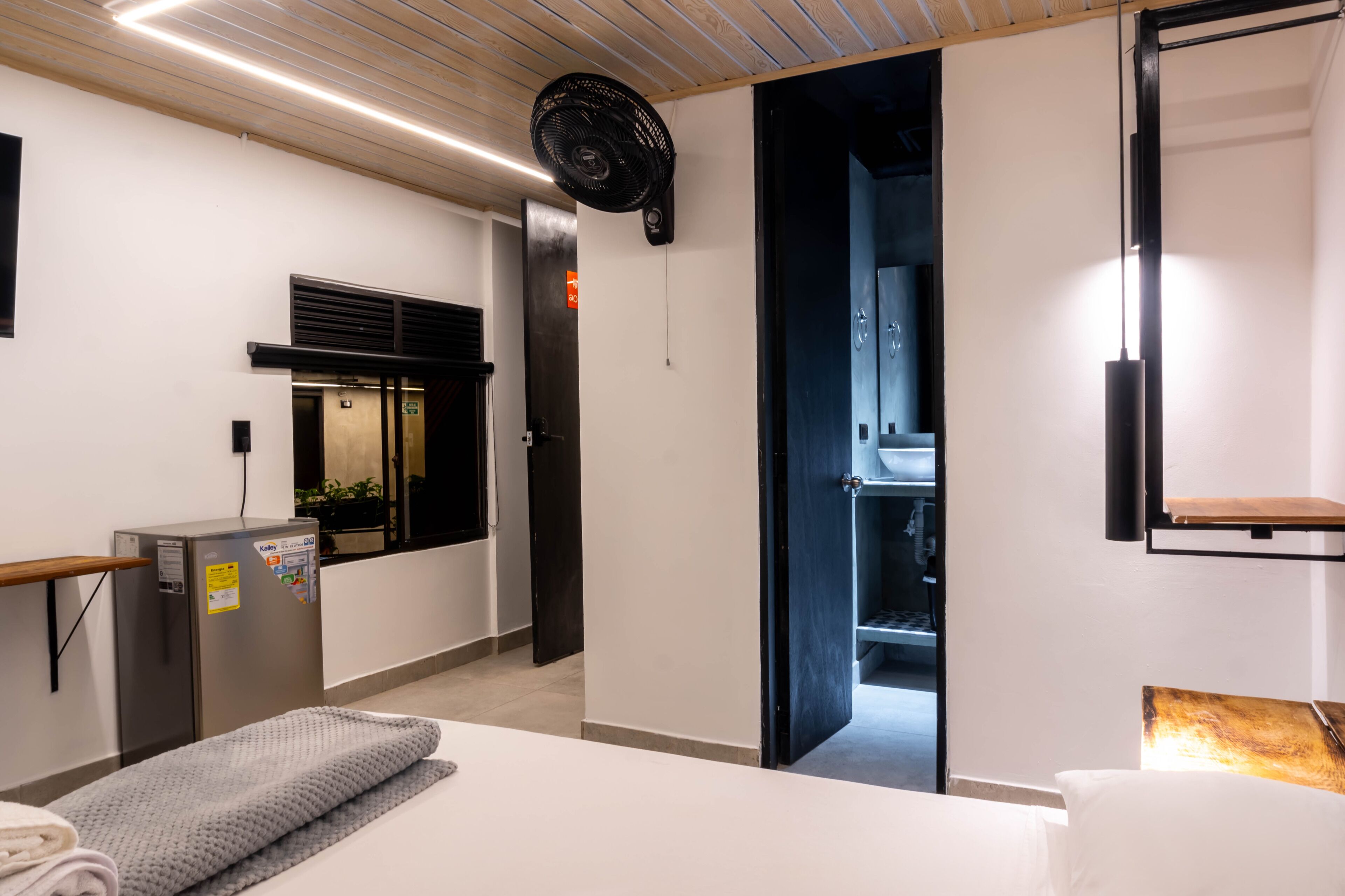 A modern hotel room with a bed, a desk, a refrigerator, a fan, a door leading to a bathroom, and a wall-mounted shelf.
