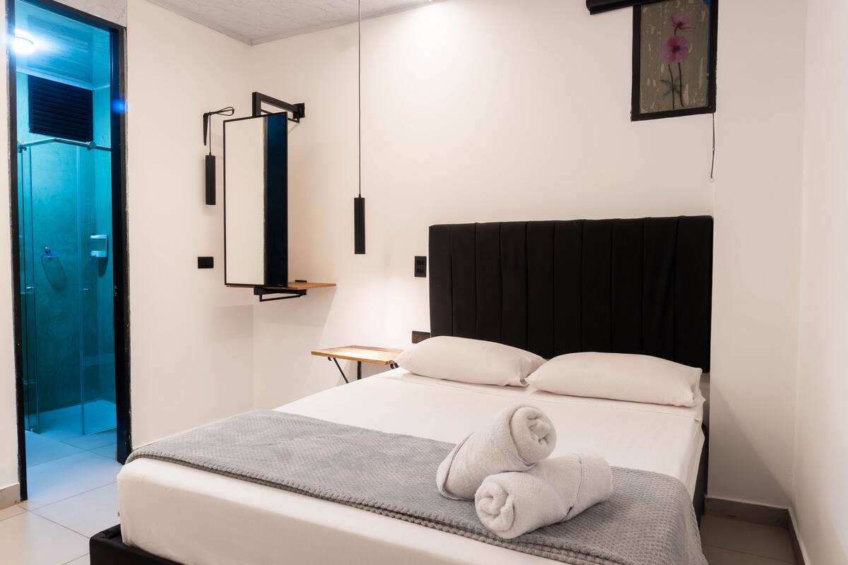 A hotel room with a bed, a bedside table, a mirror, a towel, and a door leading to a bathroom.