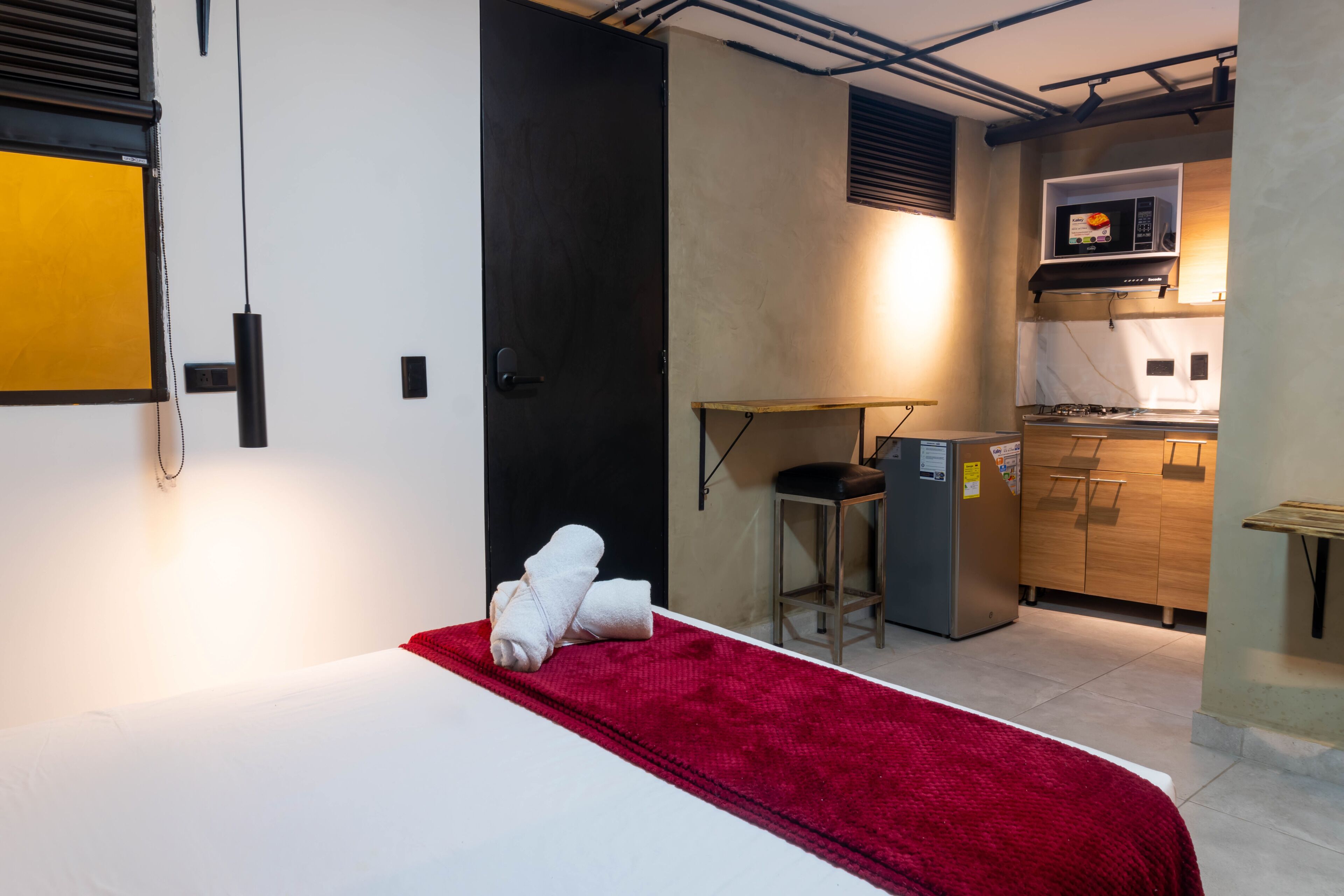 Une chambre d’hôtel compacte comprenant un lit, un plaid rouge, une petite kitchenette équipée d’un four à micro-ondes et une porte donnant sur une autre pièce.
