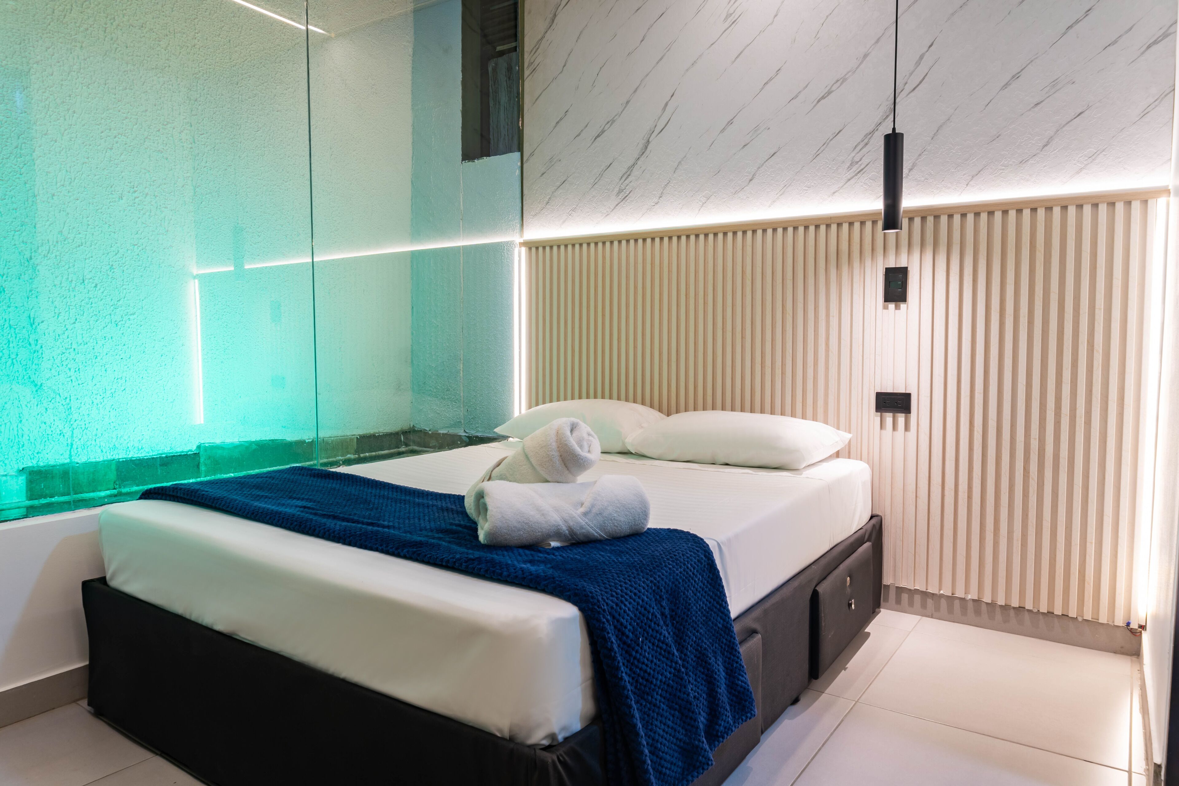 Quarto moderno com divisória de vidro, cama com roupa de cama branca e manta azul, e parede de ripas de madeira.
