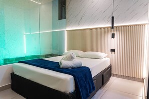 Un dormitorio moderno con separador de vidrio, cama con ropa de cama blanca, manta azul y pared de listones de madera.