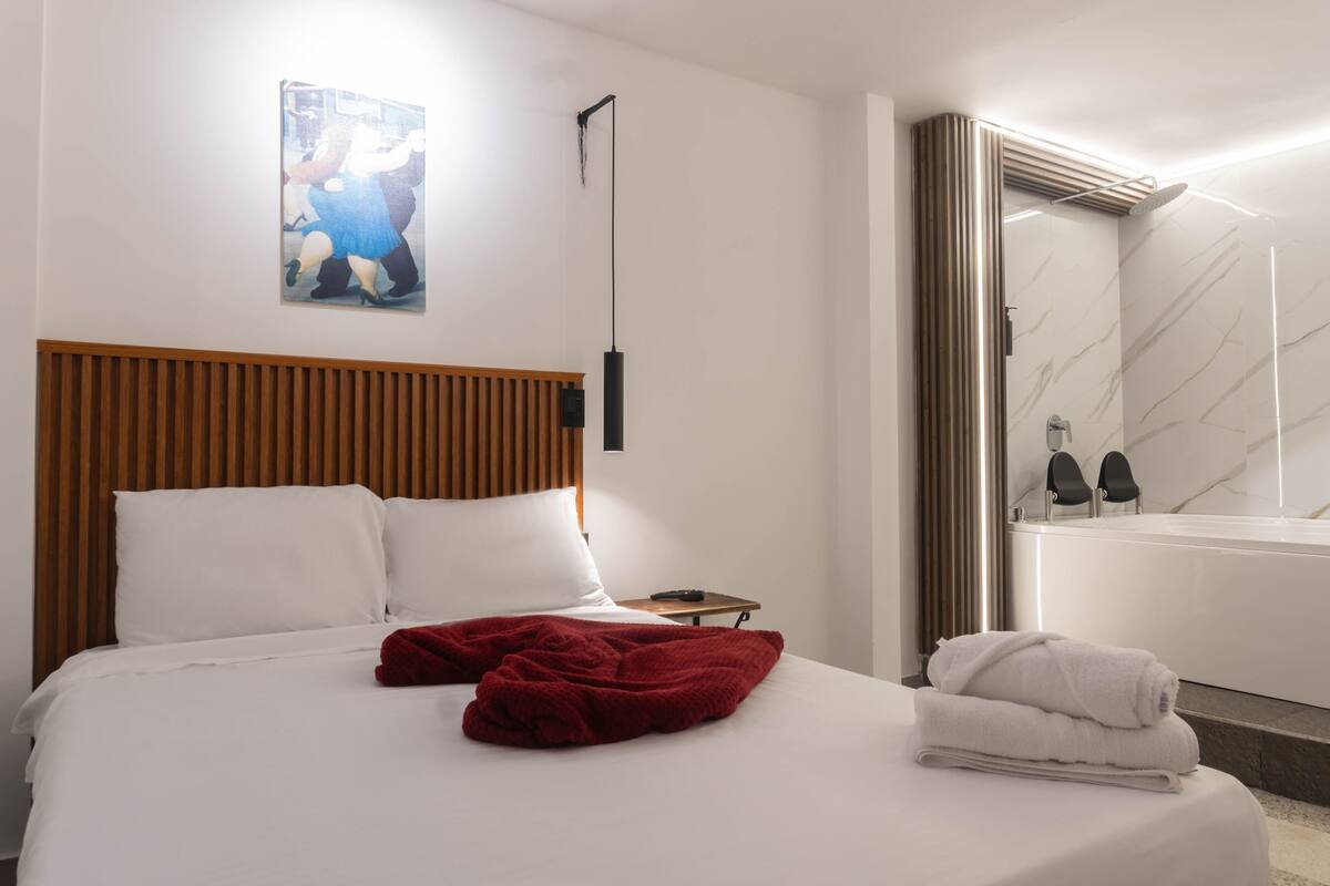 A hotel room with a bed, a nightstand, a painting, a bathroom with a bathtub, and a shower.