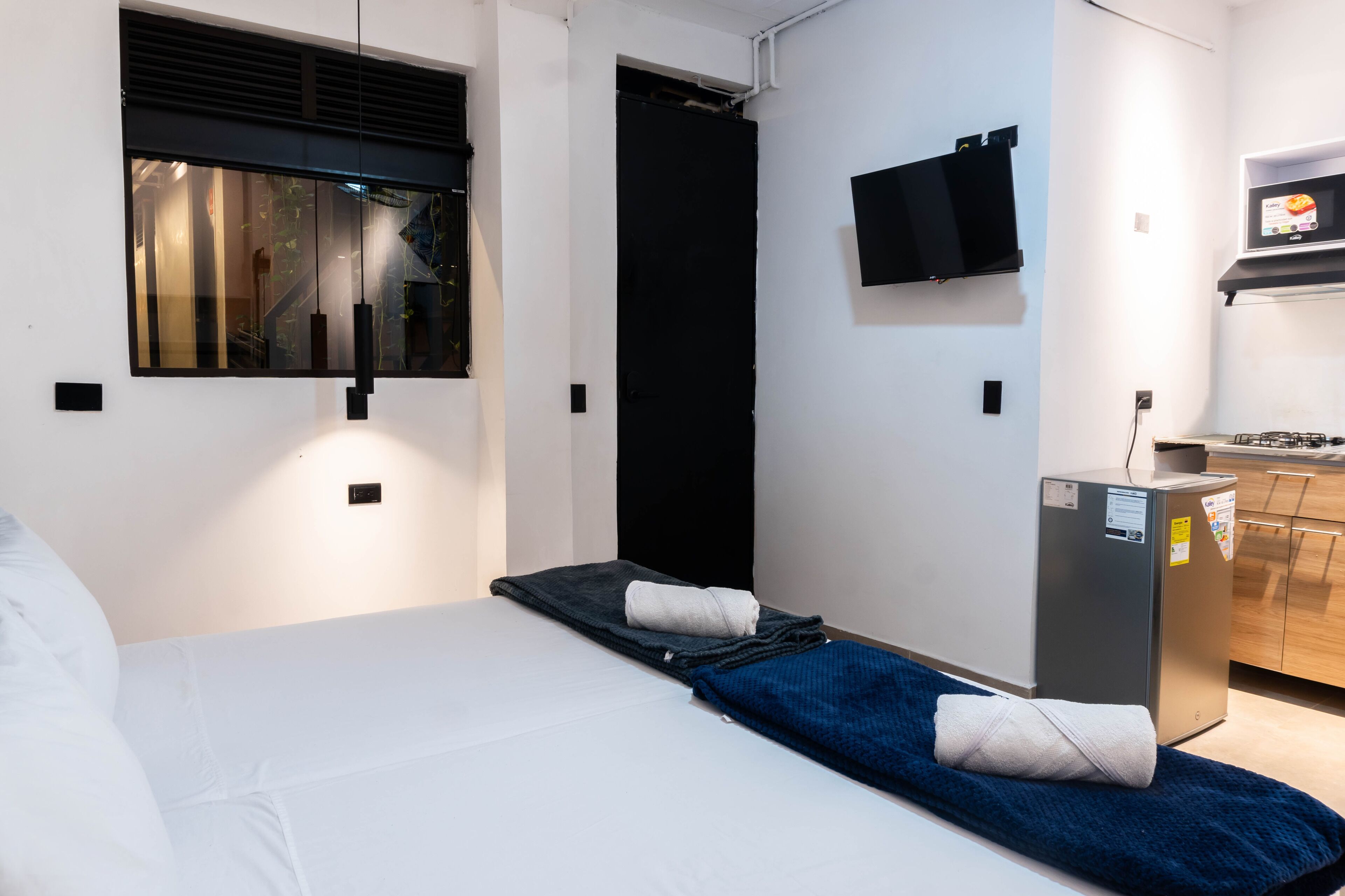 A hotel room with a bed, a small desk with a computer, a window with blinds, and a kitchen area with a refrigerator.