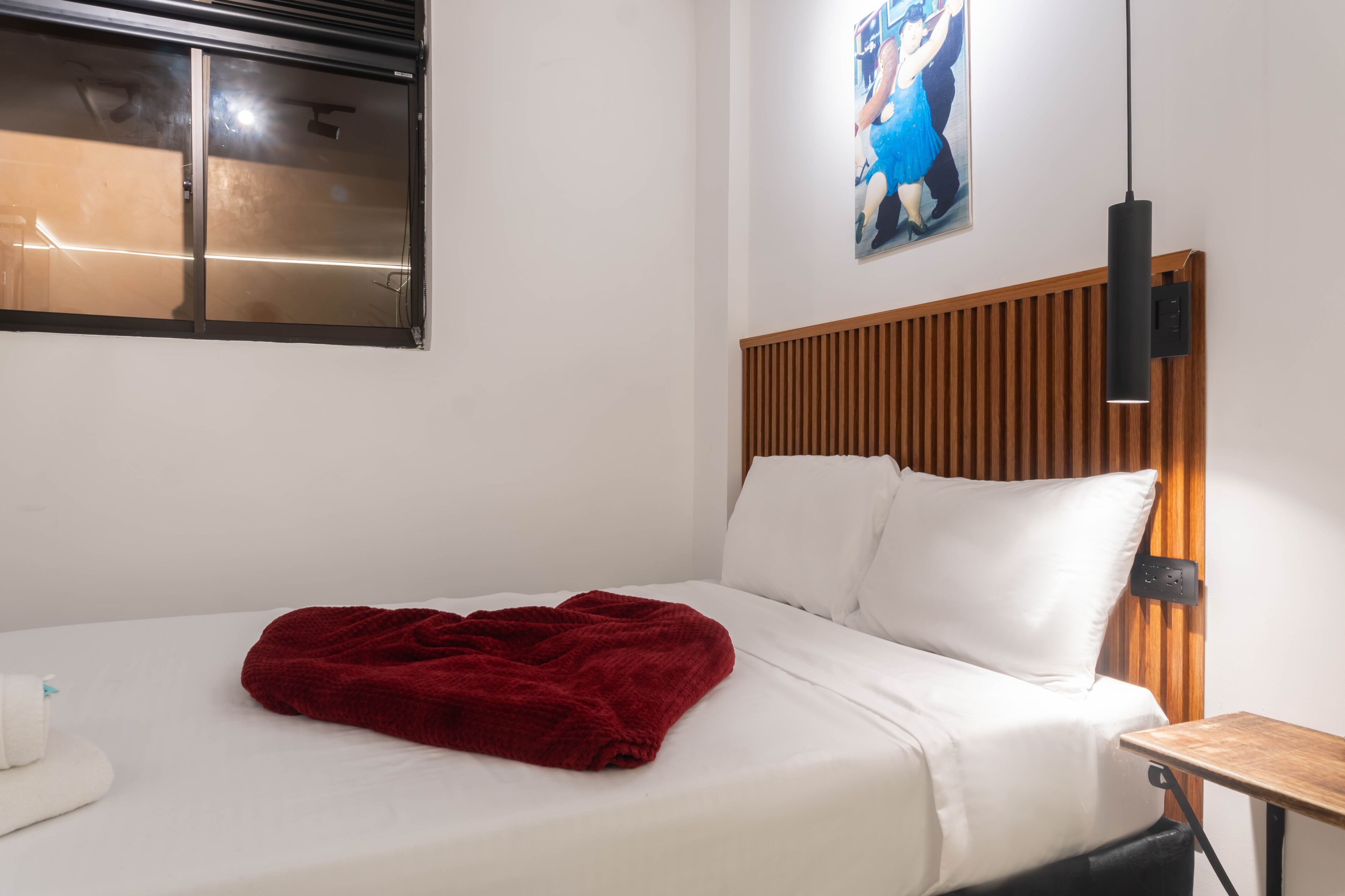 A bed with white bedding and a red blanket, a wooden headboard, a wall-mounted light, and a painting above the bed.