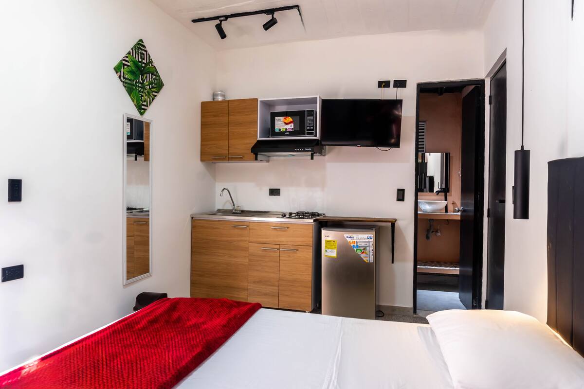 A compact hotel room with a kitchenette, a bed, a red throw blanket, and a mirror.