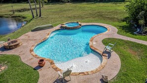 Una piscina con un jacuzzi, rodeada por un jardín ajardinado con un estanque pequeño y un pozo de fuego.