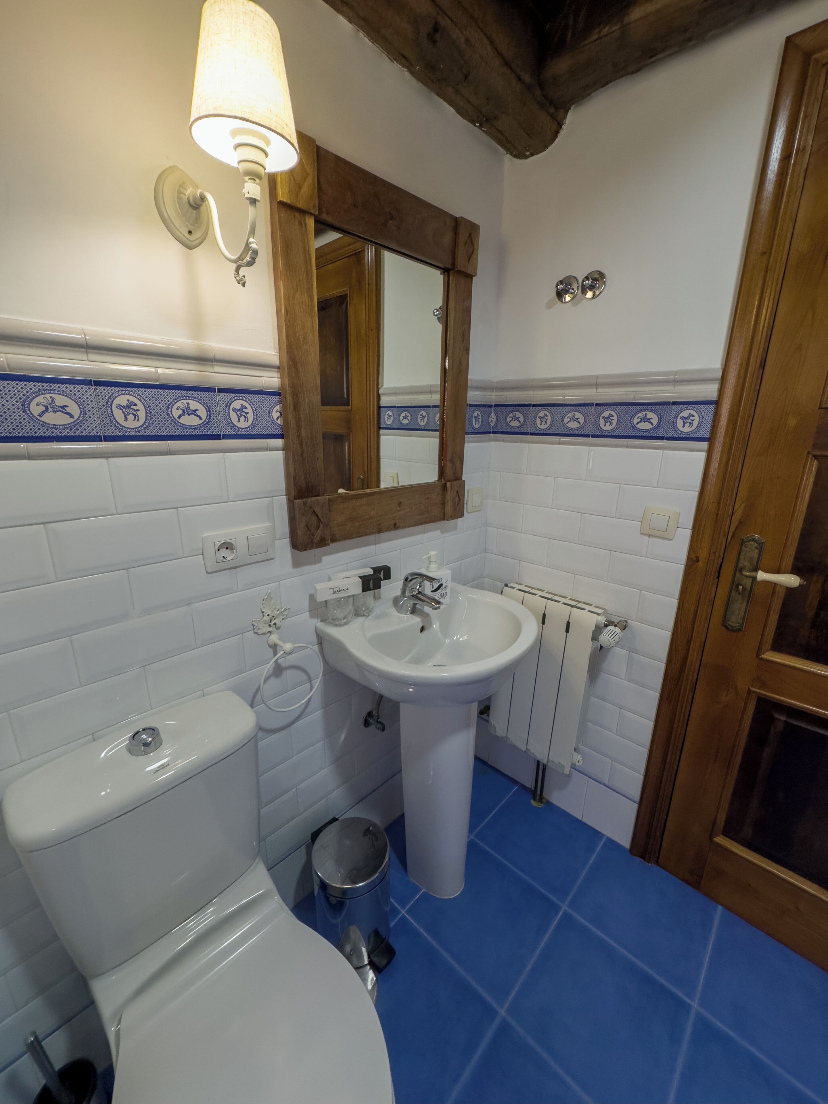 A bathroom with a white toilet, a sink with a mirror above it, a blue tiled floor, and a wooden ceiling beam.