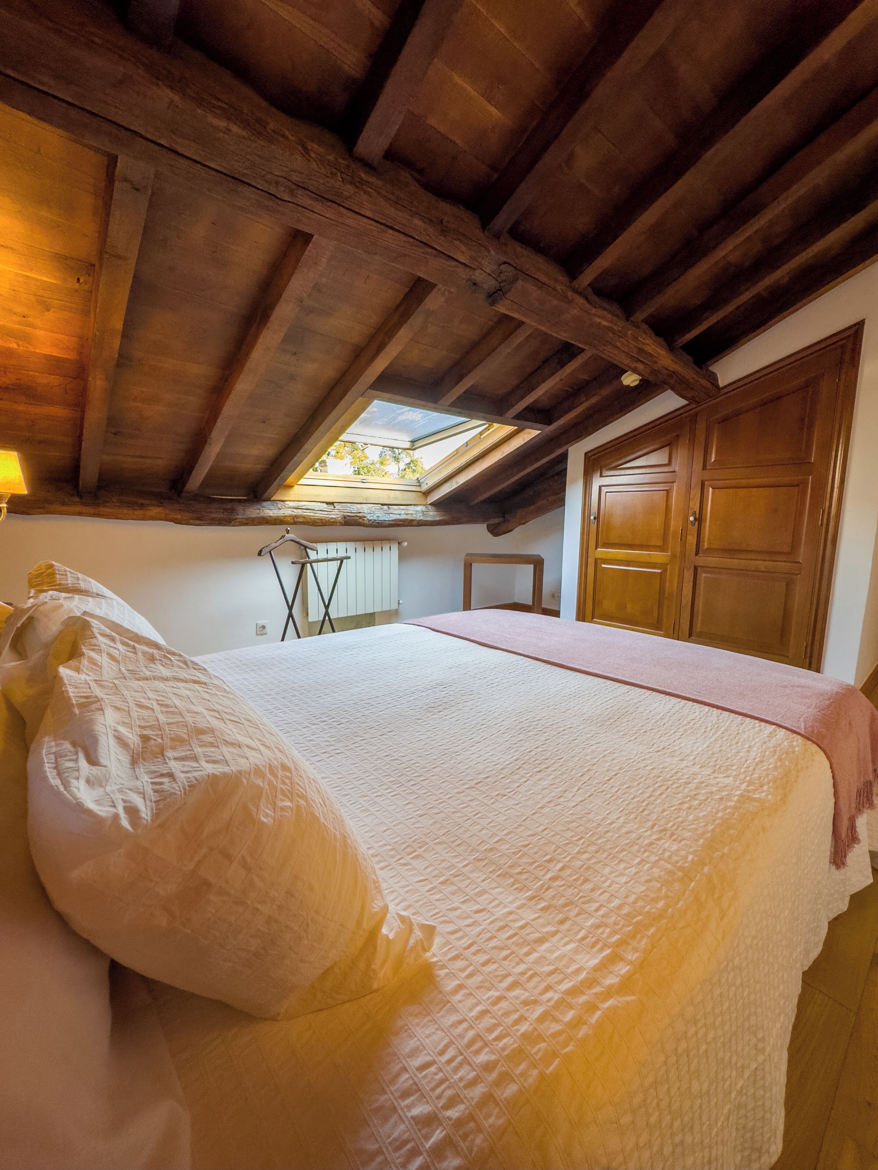 Une chambre avec un plafond en pente, un puits de lumière, un lit recouvert d’une courtepointe matelassée, une porte en bois et un radiateur.