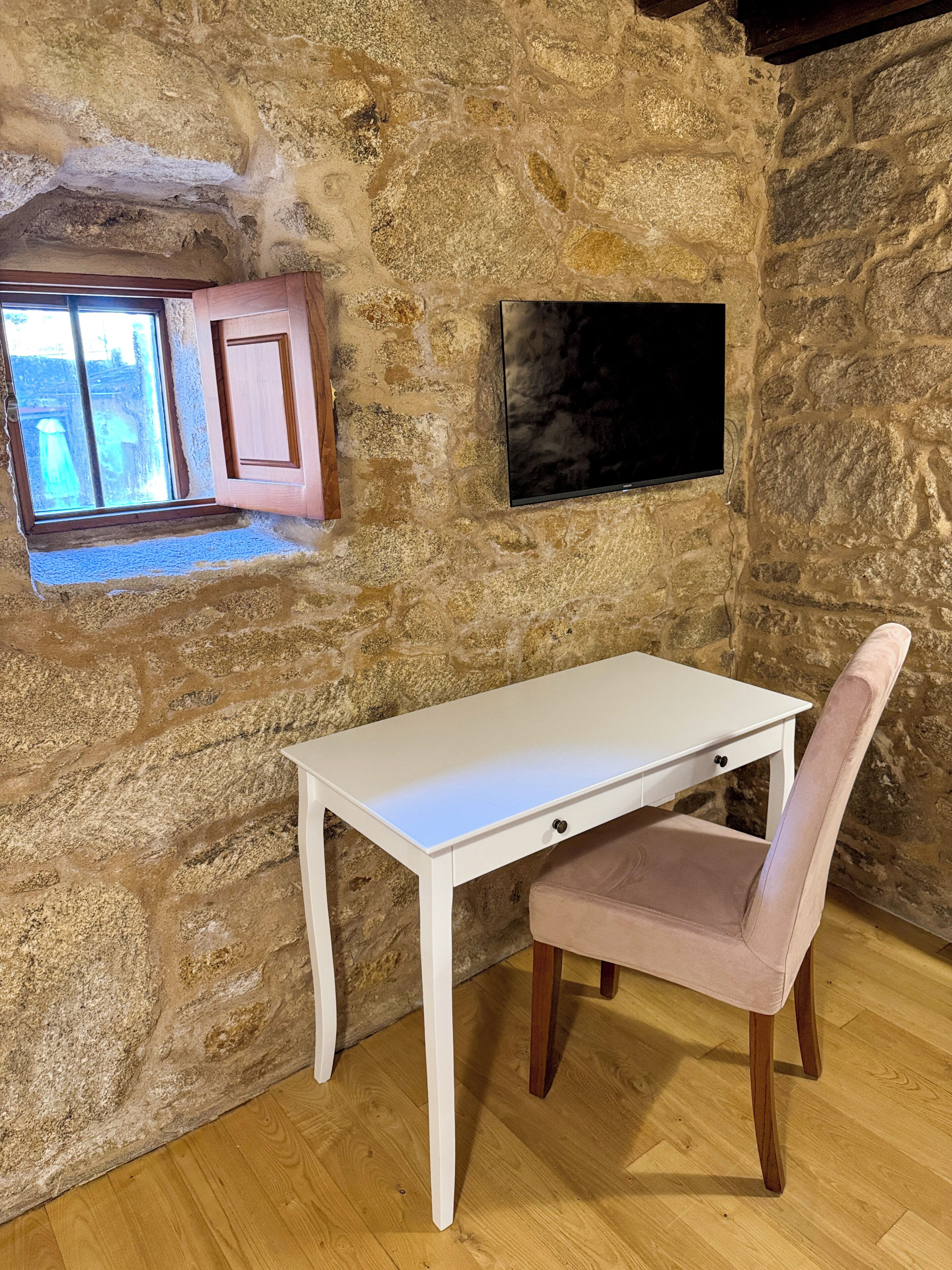 Una habitación con paredes de piedra, un escritorio blanco y una silla, un ventanal pequeño y un cuadro con marco negro en la pared.