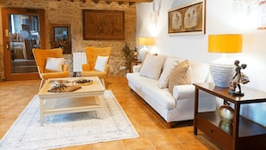 A cozy living room with a stone wall, wooden beams, a white sofa, a yellow armchair, a coffee table, and a small side table with a lamp.