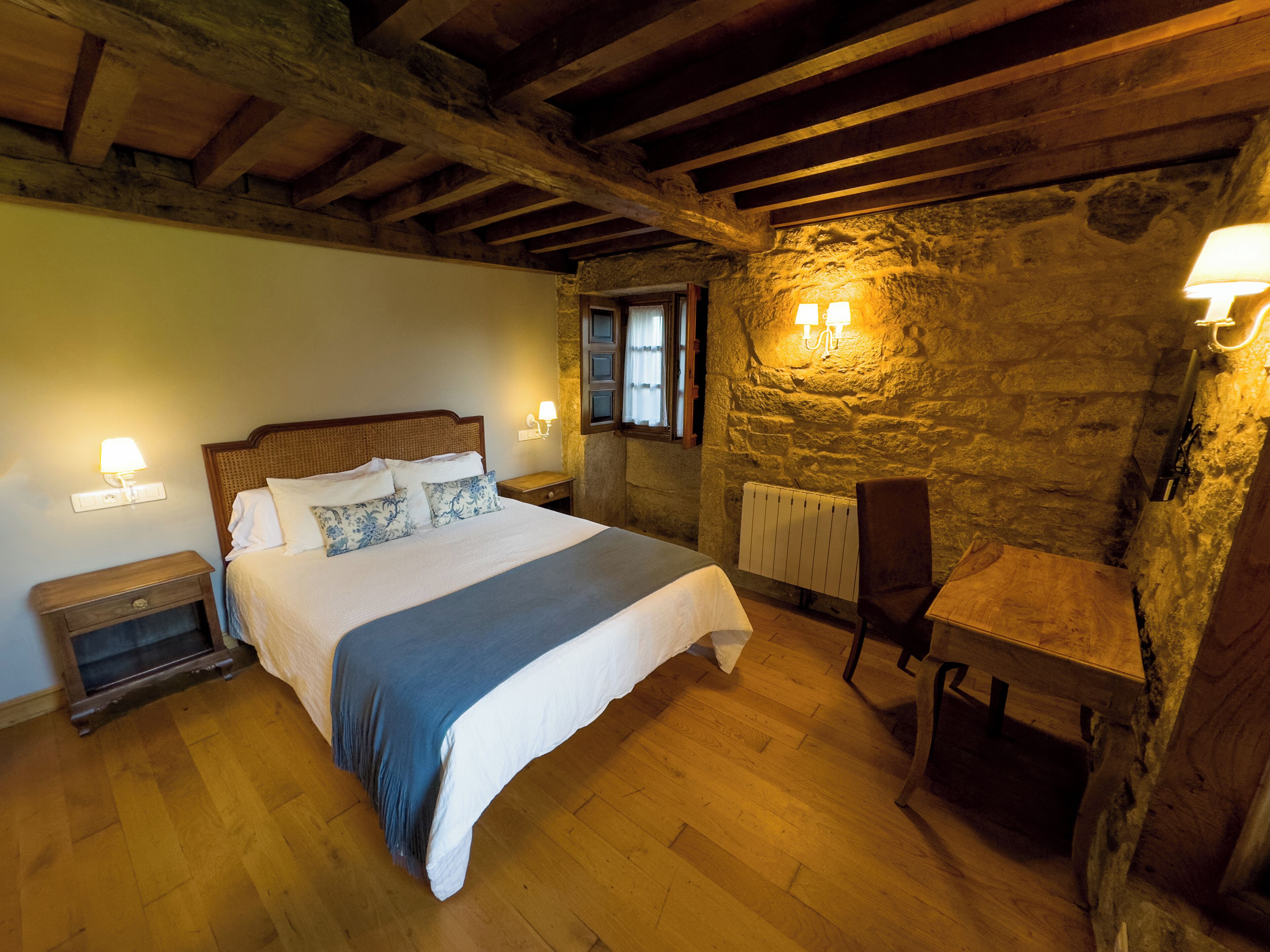 A bedroom with a wooden bed, a nightstand, a chair, a stone wall, and a window.
