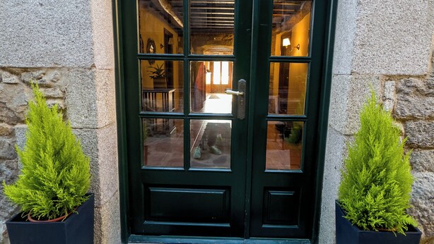 A stone building entrance with a dark green door, two potted plants, and a blue mat at the entrance.