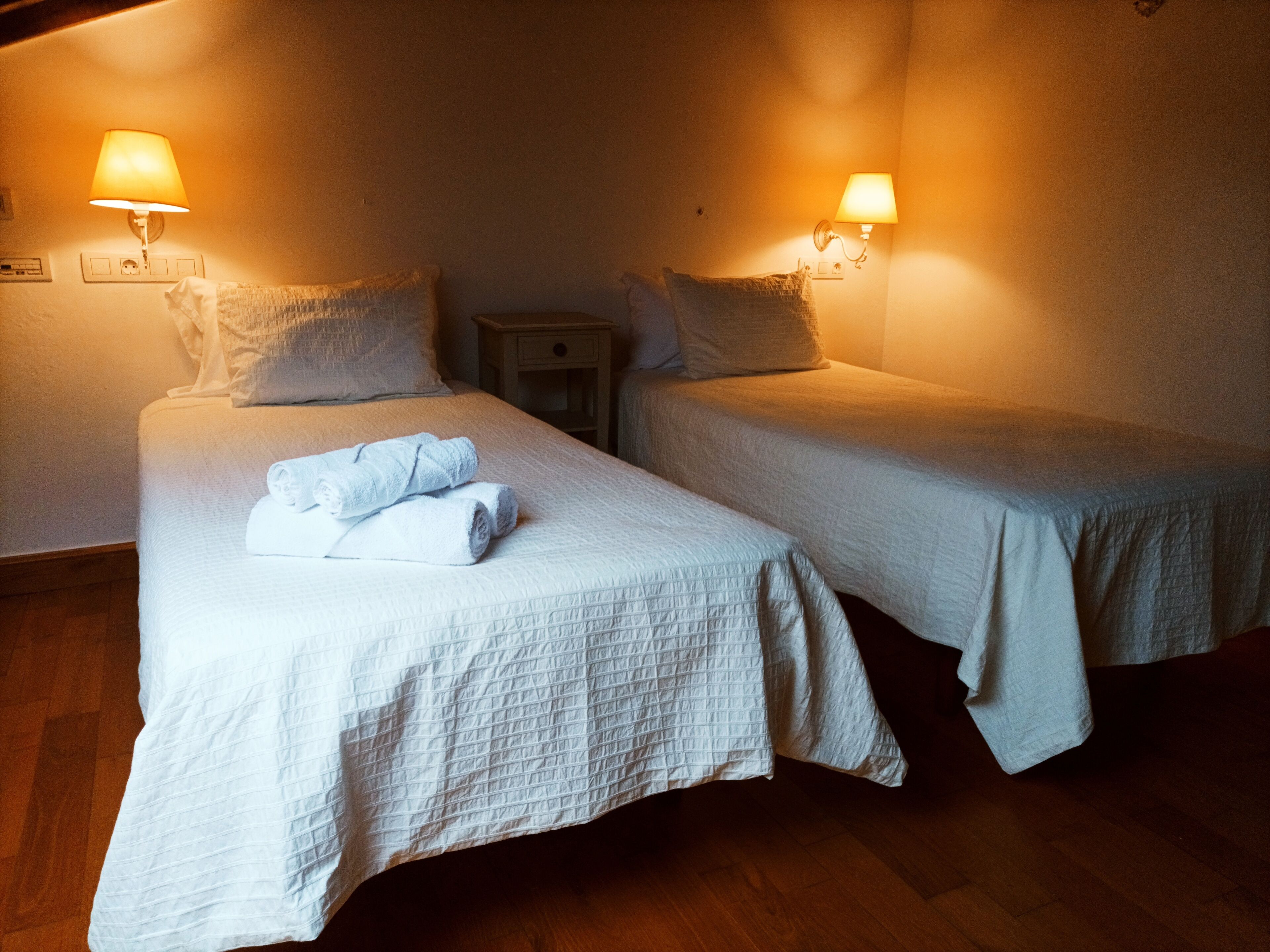 Two single beds with white linens, a wooden nightstand, and wall-mounted lamps.