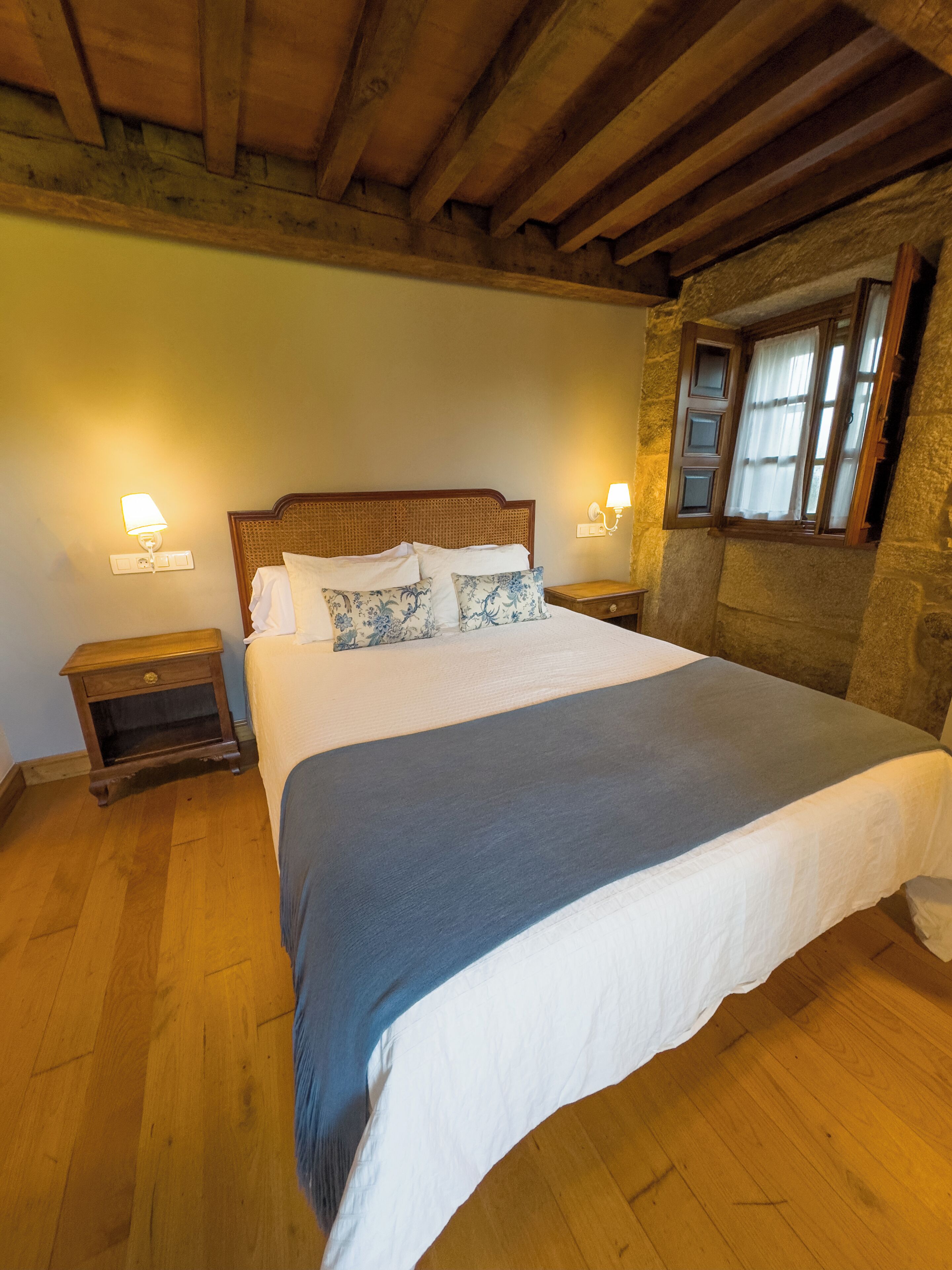A bedroom with a wooden ceiling, a large bed with a blue blanket, two bedside tables with lamps, and a stone wall.