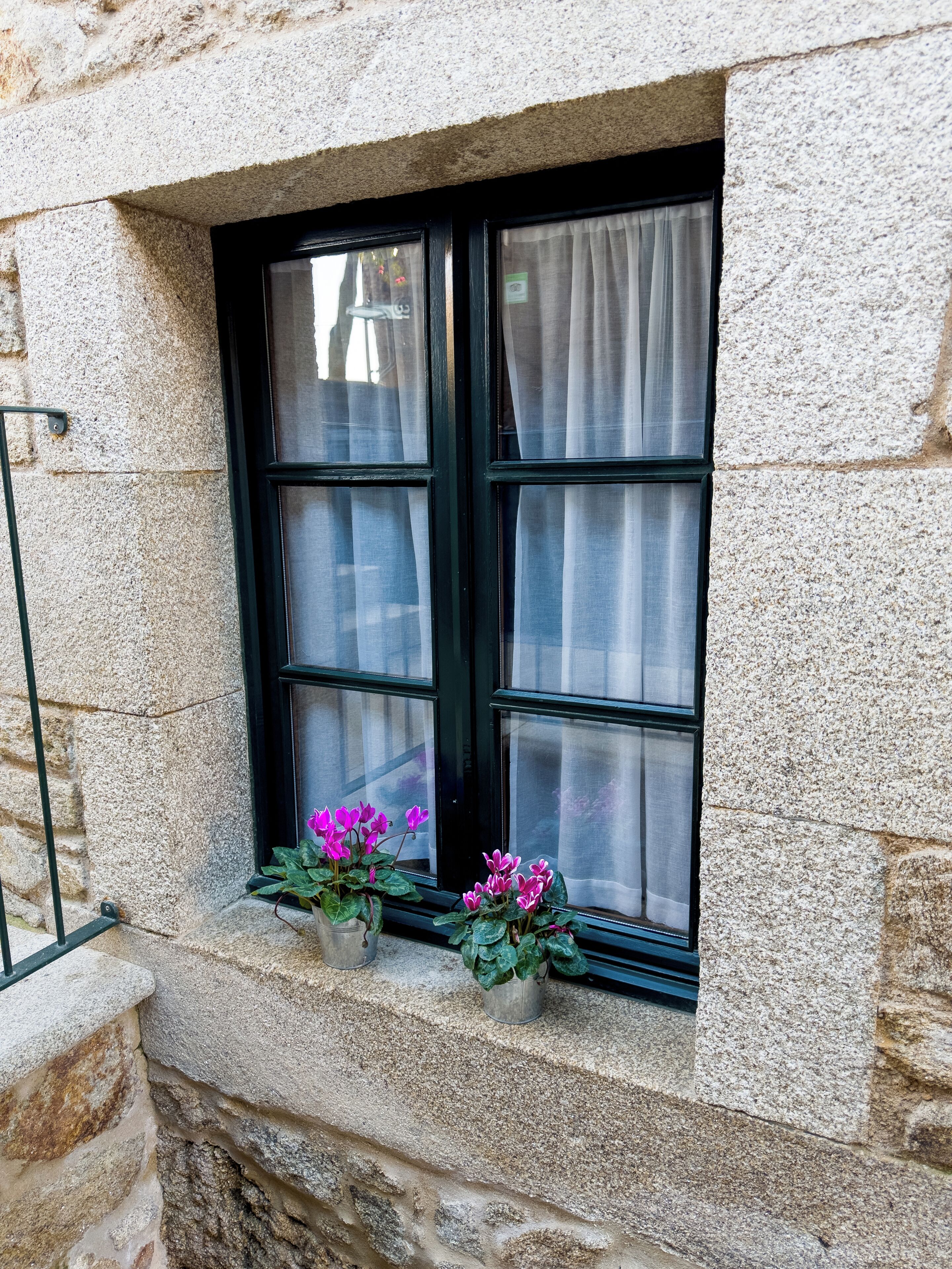 Un edificio de piedra con una ventana de marco negro y cortinas blancas. Hay dos macetas con flores rosadas en el alféizar de la ventana.