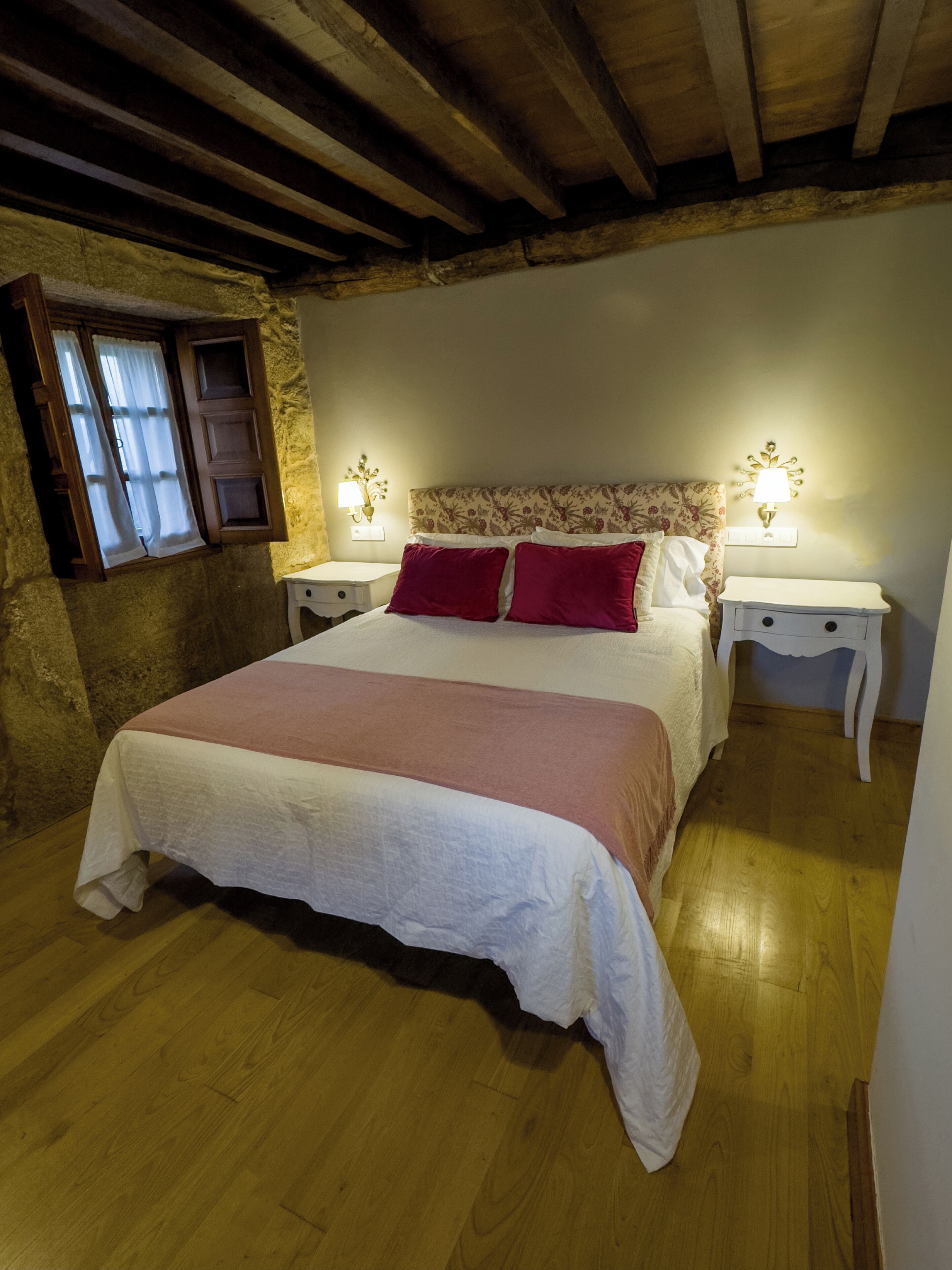 Une chambre avec un plafond en bois, un lit recouvert d’une couvre-lit blanche et rose, deux tables de chevet avec des lampes et une fenêtre avec des rideaux.