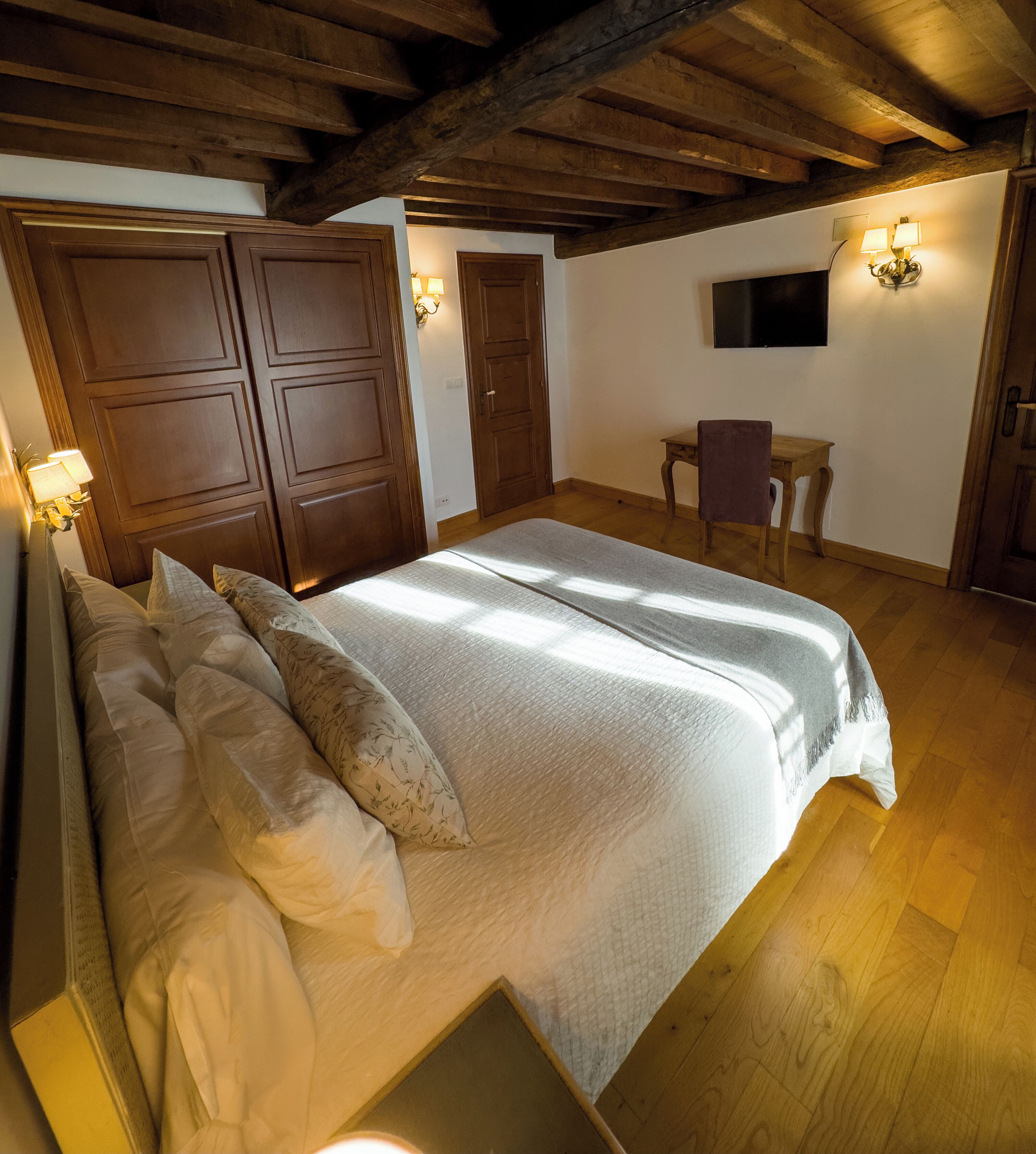Un dormitorio con una cama grande, vigas de madera en el techo, un escritorio con una silla y un televisor montado en la pared.